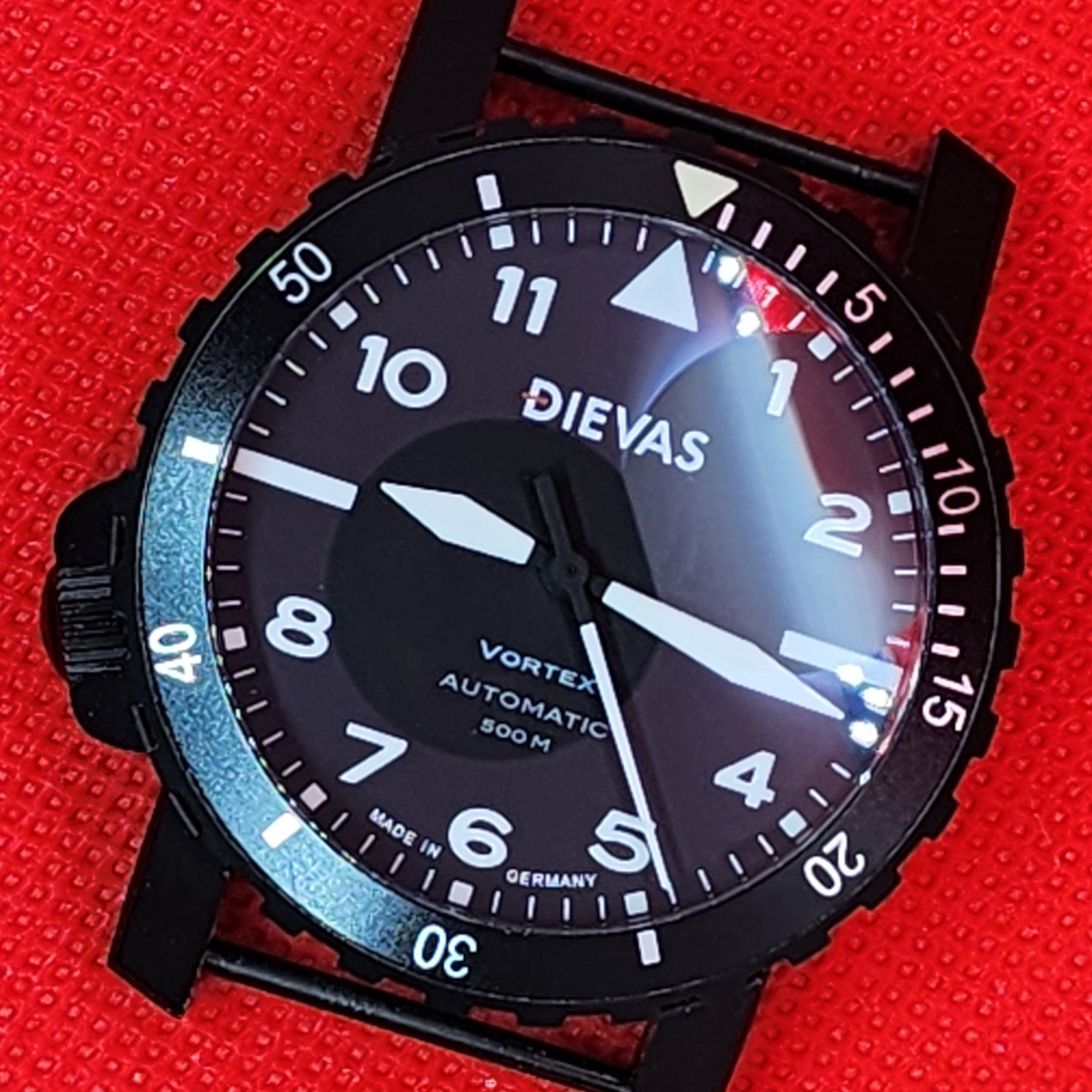 A black Dievas Vortex automatic watch with a black dial and bezel, featuring white Arabic numerals and hands, resting on a textured red background.