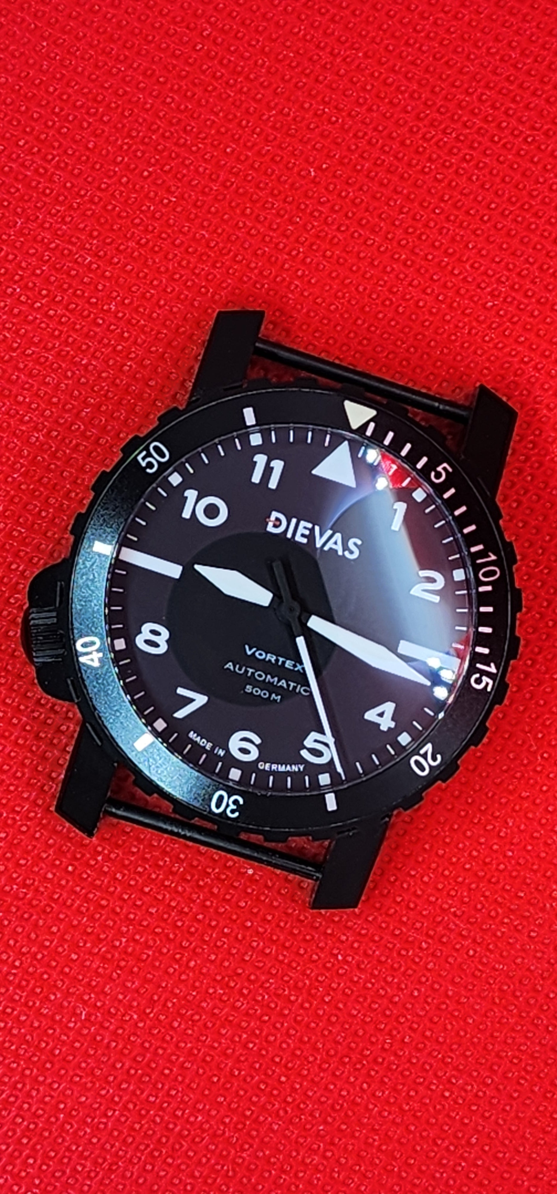 A black Dievas Vortex automatic watch with a black dial and bezel, featuring white Arabic numerals and hands, resting on a textured red background.