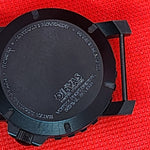 Black round watch case back with a visible 'DIEVAS' logo, photographed on a textured red background.
