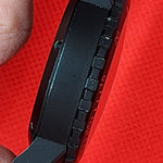 Close-up of a black tactical watch case held between fingers against a bright red textured background.