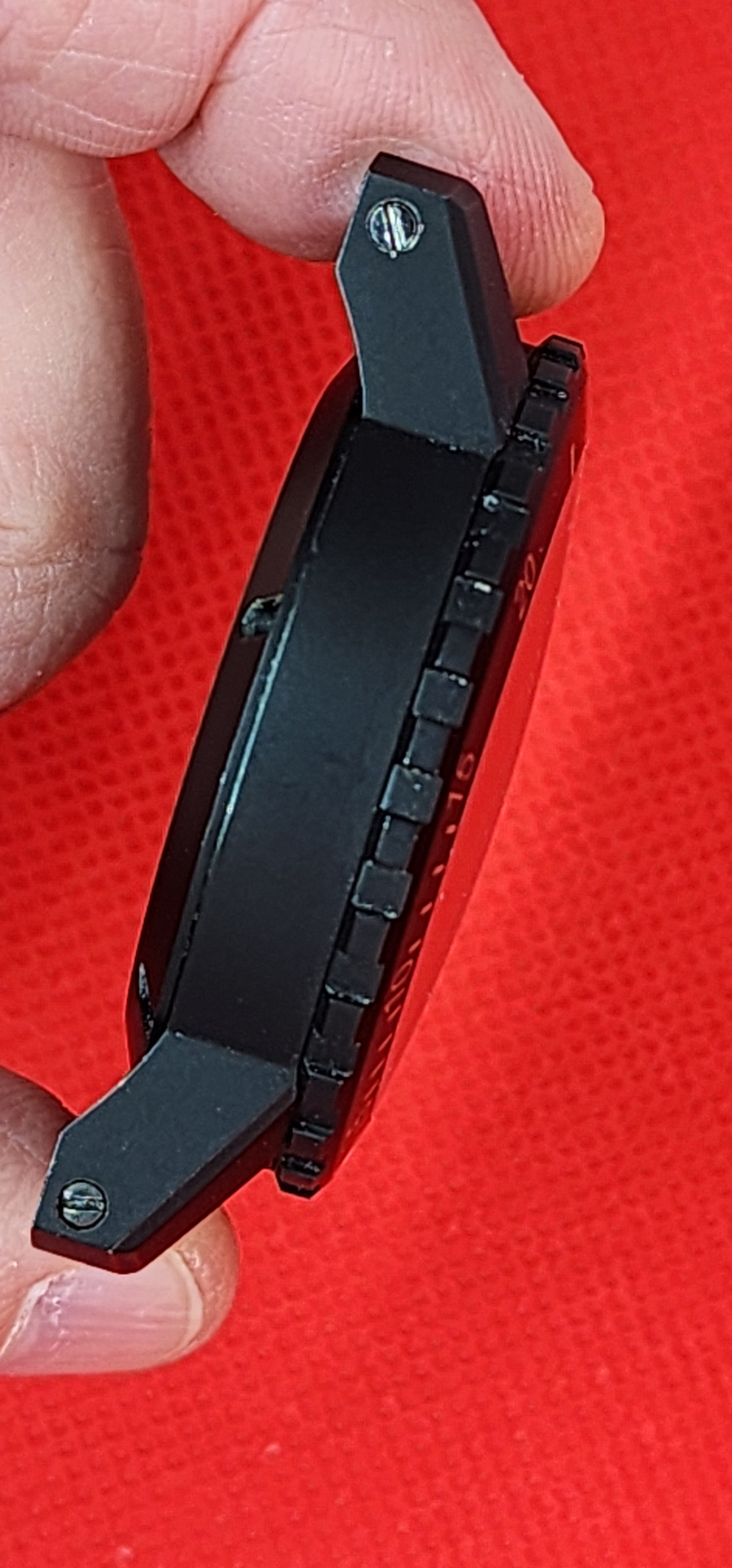 Close-up of a black tactical watch case held between fingers against a bright red textured background.
