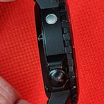 A close-up of a black tactical professional vortex watch side profile being held between fingers against a bright red textured background.
