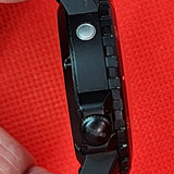 A close-up of a black tactical professional vortex watch side profile being held between fingers against a bright red textured background.