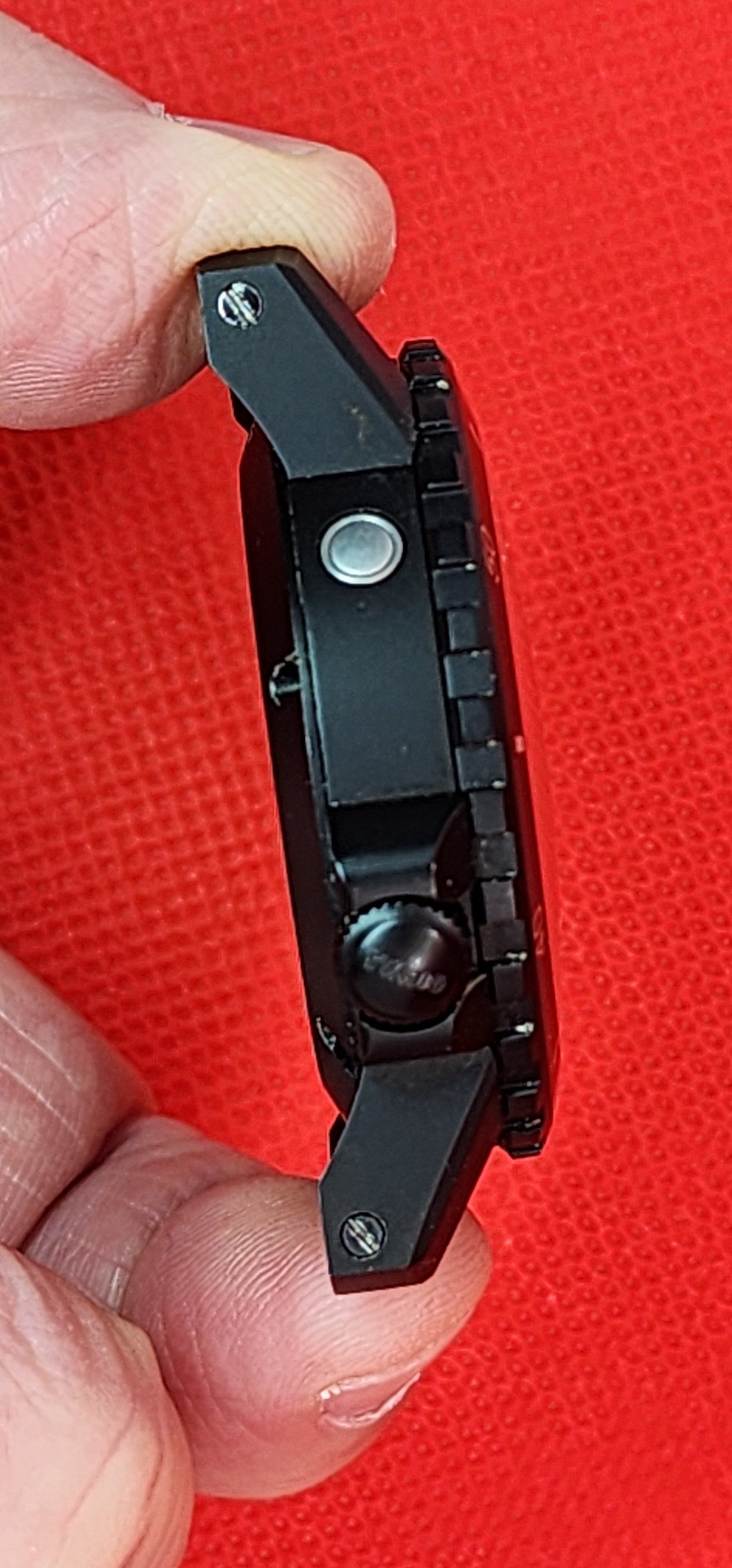 A close-up of a black tactical professional vortex watch side profile being held between fingers against a bright red textured background.