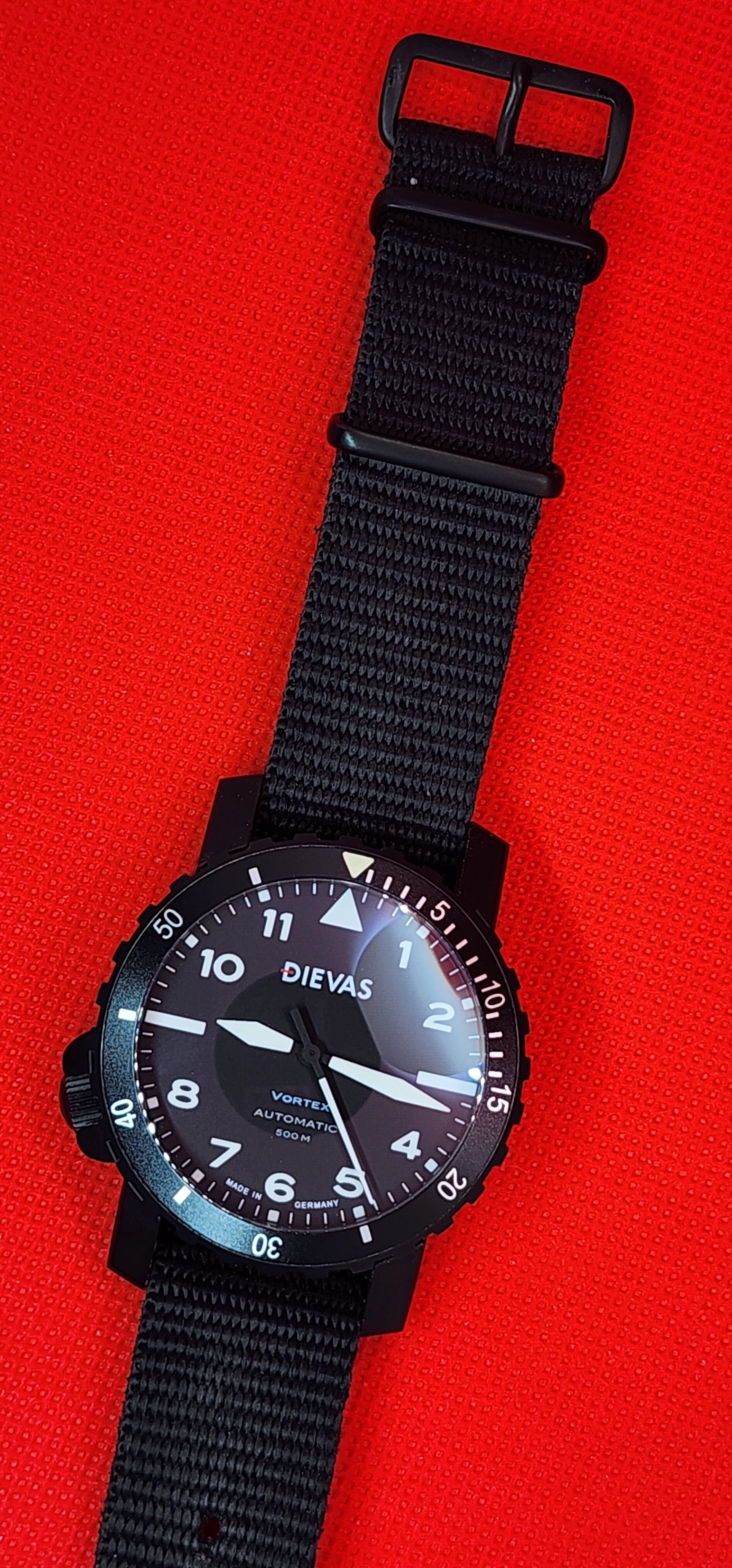 A black dial watch with a black fabric NATO-style strap, photographed on a bright red textured background. The brand name 'DIEVAS' is visible on the watch dial.

