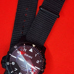 A black dial-style wristwatch with a black fabric NATO-style strap, photographed on a bright red textured background.