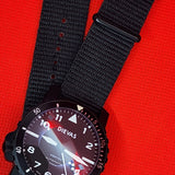 A black dial-style wristwatch with a black fabric NATO-style strap, photographed on a bright red textured background.