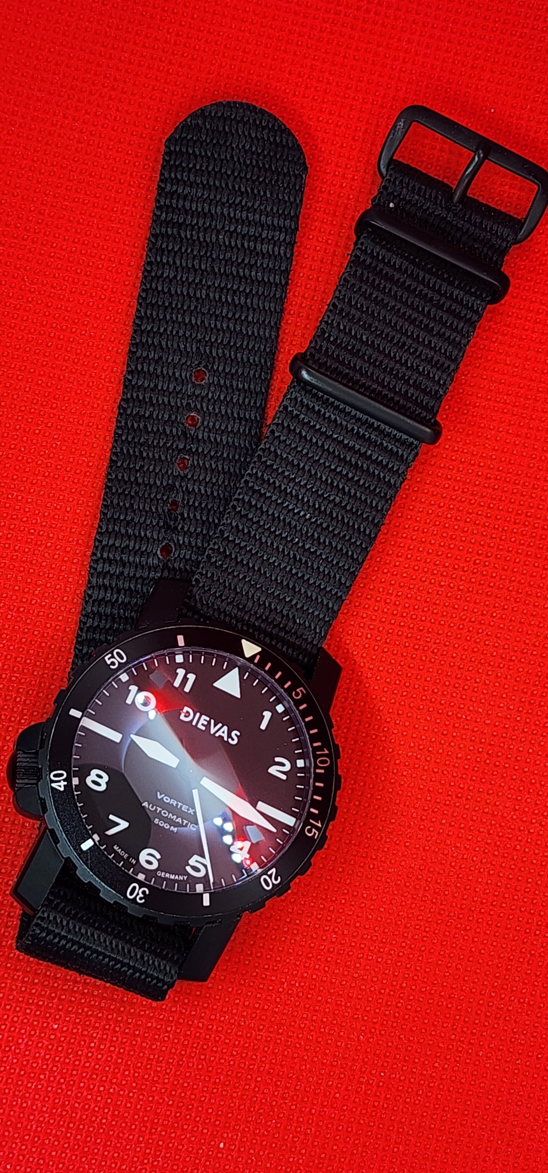 A black dial-style wristwatch with a black fabric NATO-style strap, photographed on a bright red textured background.